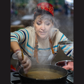 A woman wearing a red fez and apron stirs a pot in a kitchen, surrounded by ingredients and steam rising from the pot.